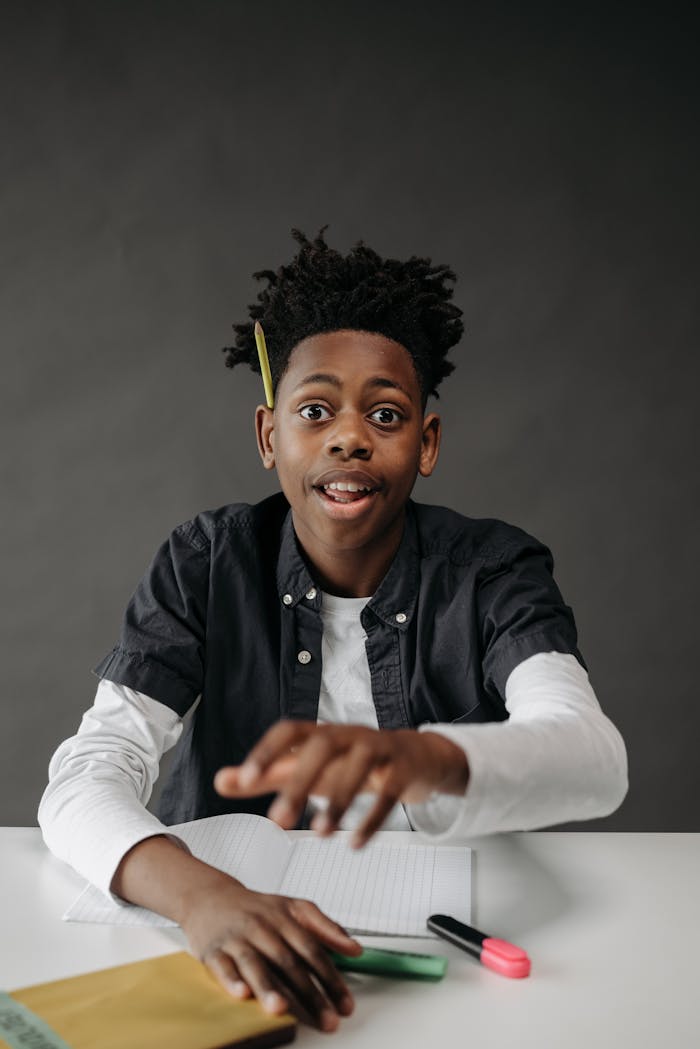 A young student with dreadlocks smiling and sitting at a desk, with a gray background.